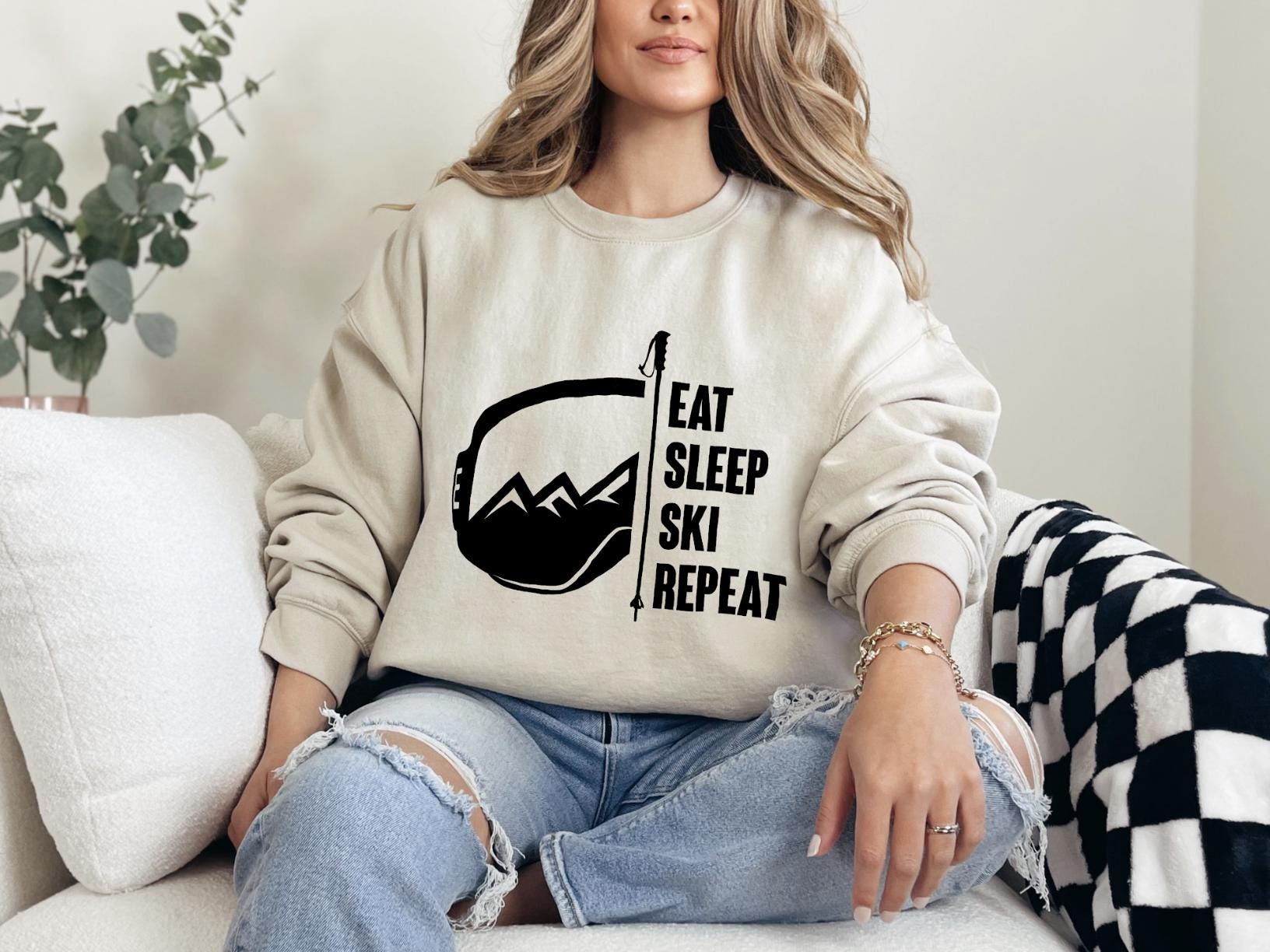 a woman sitting on a couch wearing a sweatshirt that says eat sleep ski repeat
