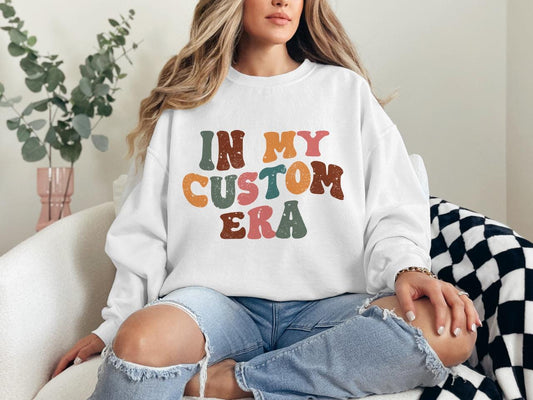 a woman sitting on a couch wearing a sweatshirt that says in my custom era